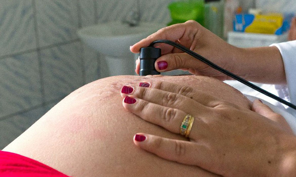 Uma mão com unhas pintadas de vinho e um anel dourado em um dos dedos em cima de uma barriga grávida. Outra mão utiliza um equipamento com fio sobre a barriga. No fundo, uma parede de azulejo e uma pia branca.