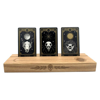 Ash wood stand featuring a hot-stamp of a skull and marigold leaves, waning crescent moon, and the sun. Excellent for holding and displaying readings while working, or for decorating your altar or desk. Comfortably fits three standard size 2.75” x 4.75” tarot or oracle cards. Stand colour may vary slightly due to differences in wood colouration. Dimensions: Approx 11.75" L x 3.5" W x 1.25" H / 30cm L x 9cm W x 3.18cm H. Composition: Ash wood. Tarot, altar, altar stand, tarot card stand, altar decor