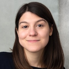 A young white woman with dark hair smiling in front of a grey backdrop.