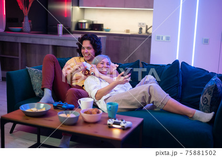 Happy boyfriend and girlfriend playing video games and holding joysticks at home. Millennial couple enjoying game while sitting on sofa and spending free time. Concept of relationship 75881502