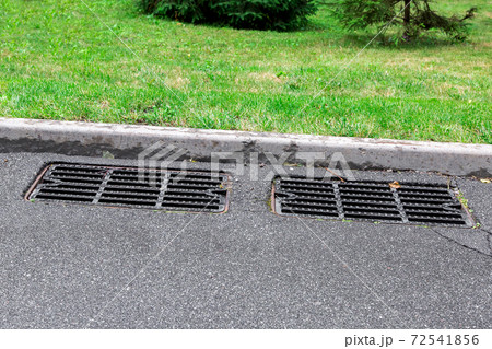 two manhole cover rain grate on an asphalt road near a stone curb on the side road green grass on sunny summer day. 72541856