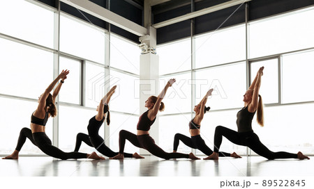 Group of young women in black sportswear doing crescent low lunge pose against panoramic window in studio 89522845