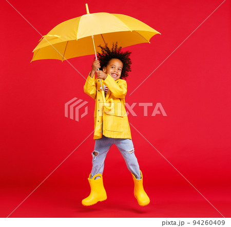 Cheerful ethnic boy jumping with umbrella against red backdrop  94260409