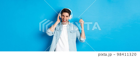 Happy smiling man enjoying listening to music in headphones, holding smartphone in raised hand, standing over blue background 98411320