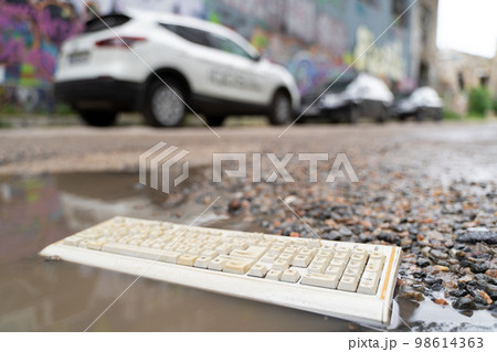 old broken computer keyboard, thrown into the trash lying on the road in a puddle. 98614363