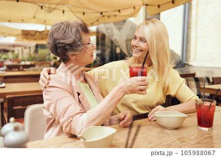 Smiling mature ladies sitting in cafe terrace talking and hugging while drinking cocktails  105988067