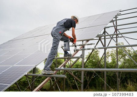 Worker fixing metal beams with electric drill for solar panels. Man wearing helmet and workwear. Photo-voltaic collection of modules as a PV panel. Array as a system of photo-voltaic panels. 107399483