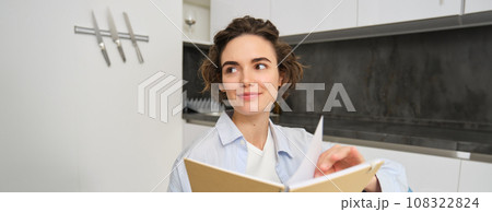 Close up portrait of modern brunette woman, holding journal, making notes in planner, reading her notebook while sitting at home in kitchen 108322824