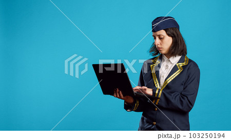 Young female airliner using laptop in studio, browsing online internet page and social media app. Air hostess working in aviation flying industry and holding portable pc, scrolling website. 103250194