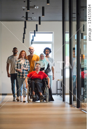 A diverse group of young business people walking a corridor in the glass-enclosed office of a modern startup, including a person in a wheelchair and a woman wearing a hijab 114155034