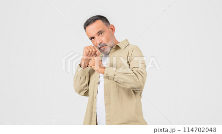 A senior man wearing a tan shirt is standing while holding his hands together in front of him in fighting pose. He appears focused and contemplative as he holds his hands clasped together. 114702048