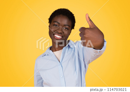 A smiling young african american woman with short hair gives a thumbs up sign against a bright yellow background, expressing positivity 114703021