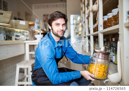 Handsome man working in package-free store using reusable containers. Zero waste shops offering package-free bulk goods and sustainable alternatives. 114726565