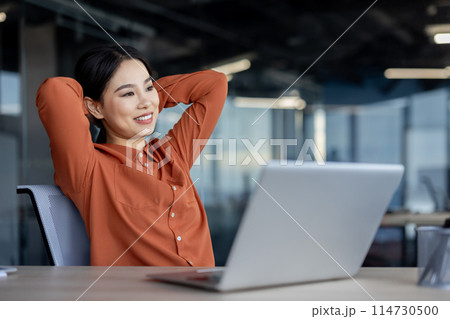 Smiling Asian businesswoman enjoying a break at the office, relaxed at her desk in a modern workspace 114730500