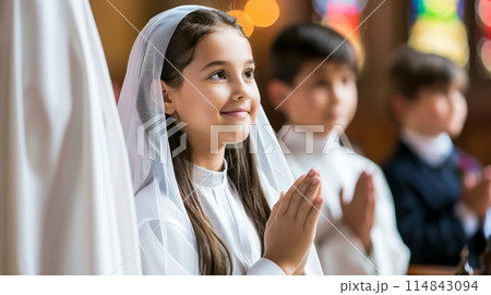 Children celebrating their First Communion in a church, dressed in white, praying, and smiling. 114843094