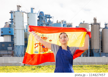 Calm young woman worker with flag of Spain against background of factory 114827457