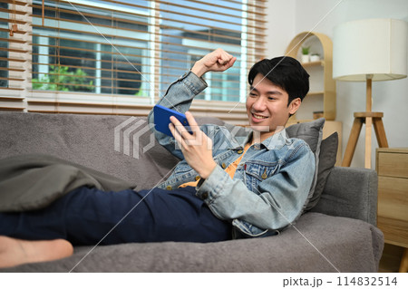 Happy young man looking at smartphone and celebrating good news sitting on couch at home 114832514