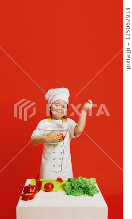 Little girl dressed as chef, holding bowl of fresh salad standing by table with vegetables, dreaming of cooking against red studio background. 115062913