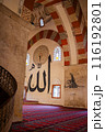 Interior of a mosque with large Arabic calligraphy on the walls and pillars with people are seen walking and praying on a red patterned carpet, in Edirne, Turkey. 116192801