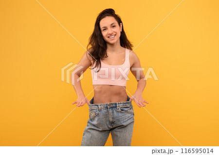 A cheerful young woman stands with hands on her hips wearing a light pink crop top and oversized blue denim jeans. She smiles confidently against a bright yellow backdrop 116595014