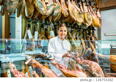 Young salesgirl in white uniform offering Iberian jamon in butcher shop 116753282