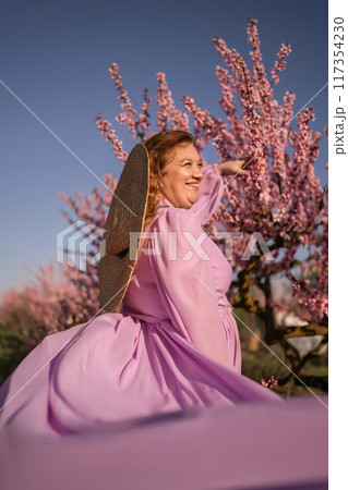 Woman blooming peach orchard. Against the backdrop of a picturesque peach orchard, a woman in a long pink dress and hat enjoys a peaceful walk in the park, surrounded by the beauty of nature. 117354230