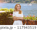 Portrait of happy blonde woman with folded arms pointing with her index finger to the right. Summer resort landscape in the background. 117943519