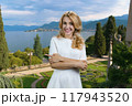 Portrait of happy blonde woman pointing with her index finger at amazing landscape. Garden with sea and mountains. 117943520