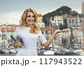 Portrait of cheerful blonde woman with tablet device shows copy space in front of a yacht docks. Vacation and tourism. 117943522