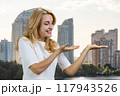 Portrait of happy mature blonde woman standing outdoors showing copy space with both hands. Residential buildings in the background. 117943526