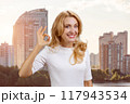 Portrait of a happy mature blond woman shows ok gesture sign. Residential area in the background. 117943534