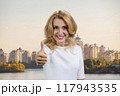 Portrait of a happy mature blond woman shows like gesture sign. Urban cityscape in the background. 117943535