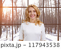 Happy mature blond woman in a white blouse standing indoors. Checkered window with winter scape view in the background. 117943538