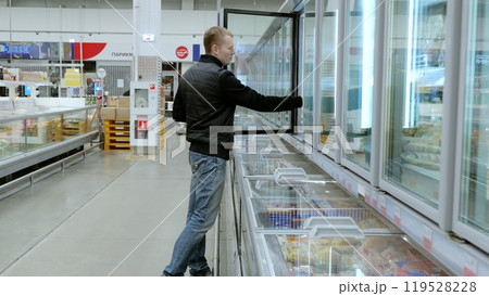 Customer browsing frozen food section in supermarket, opening freezer door to find refrigerated items. A man thoughtfully chooses products in a grocery store 119528228