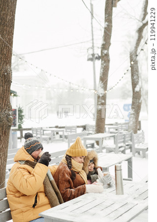 Vertical wide angle shot of modern young family sitting at outdoor cafe table together and enjoying hot drinks with snow falling 119941229