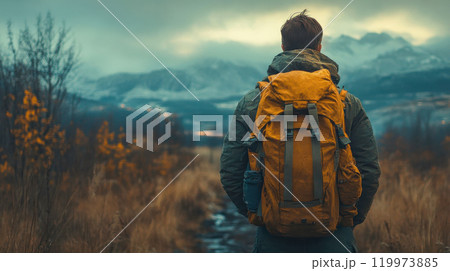 Man with yellow backpack looking towards snow-capped mountains under overcast sky. 119973885