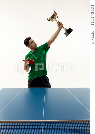 Player in victory pose holds cup and racquet at table tennis match against white studio background. Power and strength. 111348021