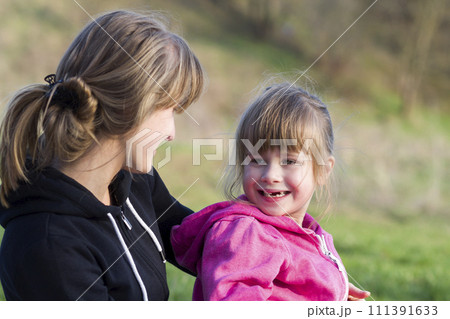 Portrait of young beautiful blond girl hugging lovingly and protectively her small preschool toothless sister, both smiling happily outdoors. Happy family relations and friendship concept. 111391633