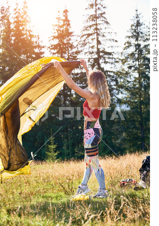 Woman traveller camping outdoors. Female tourist wearing sportswear, putting tent, making campsite in mountains. Concept of tourism and beauty of nature. 113238058