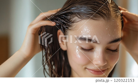 A young woman gently massages a hair treatment into her scalp, enjoying a spa-like moment in a well-lit bathroom. 120322153