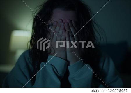 A woman with wet hair hides her face behind her hands in a dark, softly illuminated room 120538981