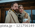 Couple bundled in thick blanket standing outdoor, hugging in front of a cabin during snowfall. 121756149