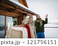 Family standing on the snowy cabin balcony, looking at beautiful winter landscape around them during snowfall. 121756151