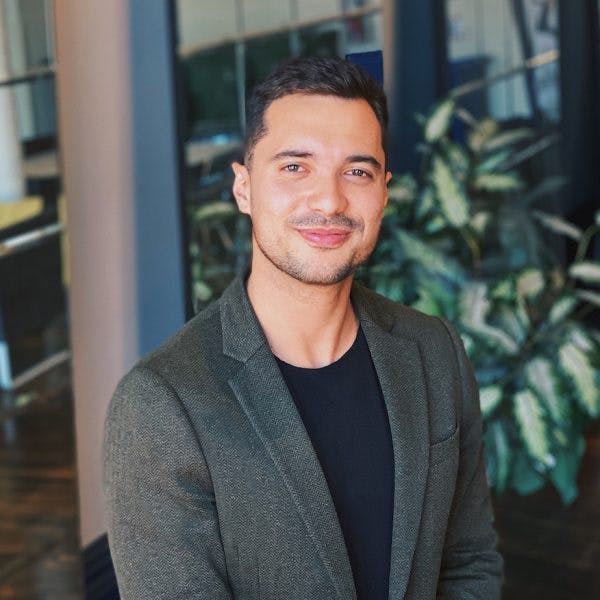 A smiling man stands in an office, wearing a dark blazer and black shirt, with plants and glass-walled rooms in the background.