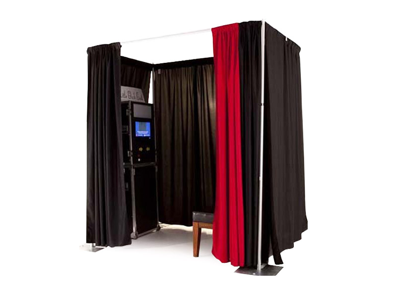 A photo booth rental set up with black pipe and drape curtains on three sides and a red curtain open on one side. There is a black photo booth box with a screen and a bench inside.