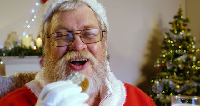 Santa Claus is savoring a cookie with a joyful expression, set against a festive background featuring a Christmas tree with lights. This image encapsulates the spirit of the holiday season, making it ideal for use in Christmas advertisements, festive greeting cards, or holiday-themed website banners.