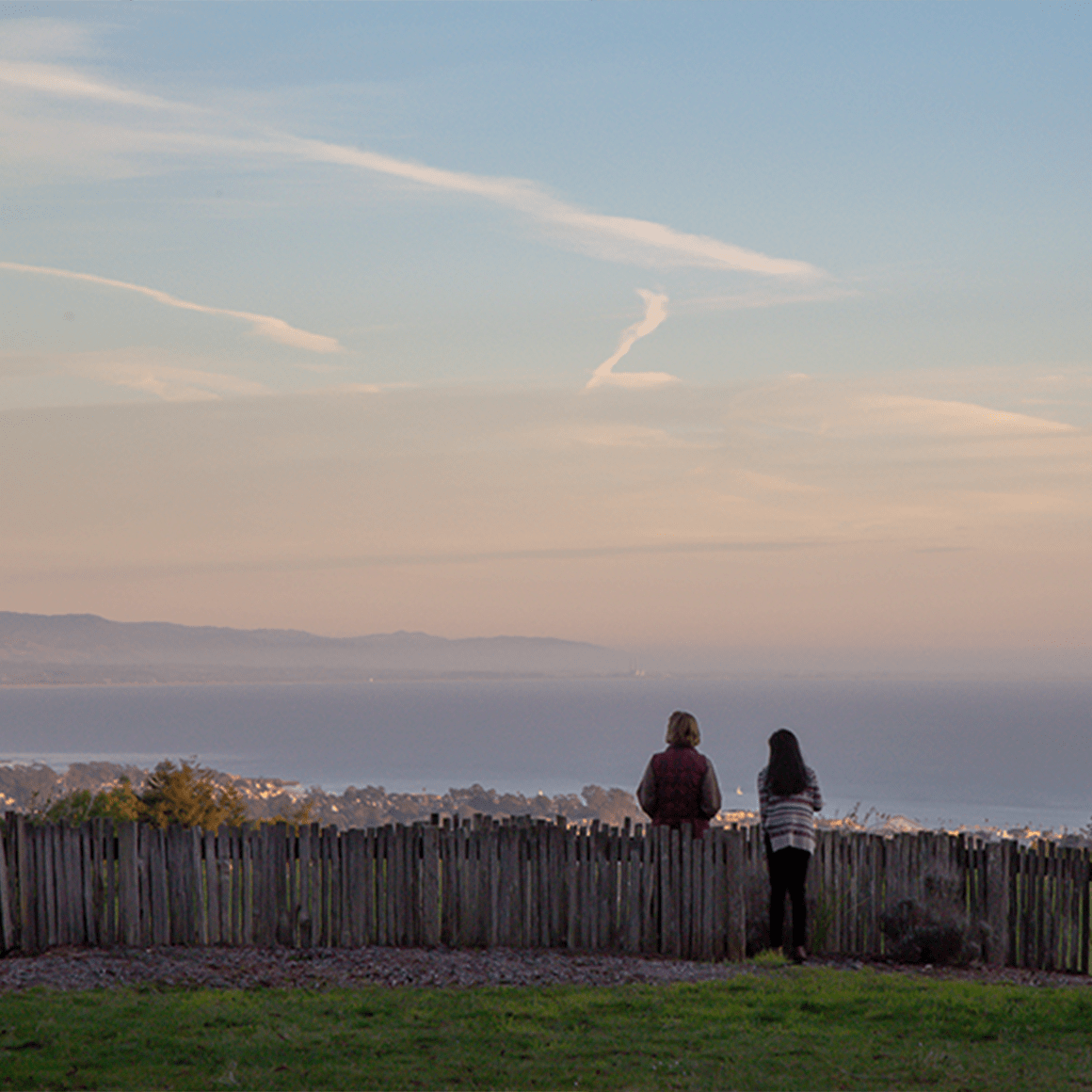 Two people, backs to camera, standing at fence on a hill, at sunset, looking at ocean. 