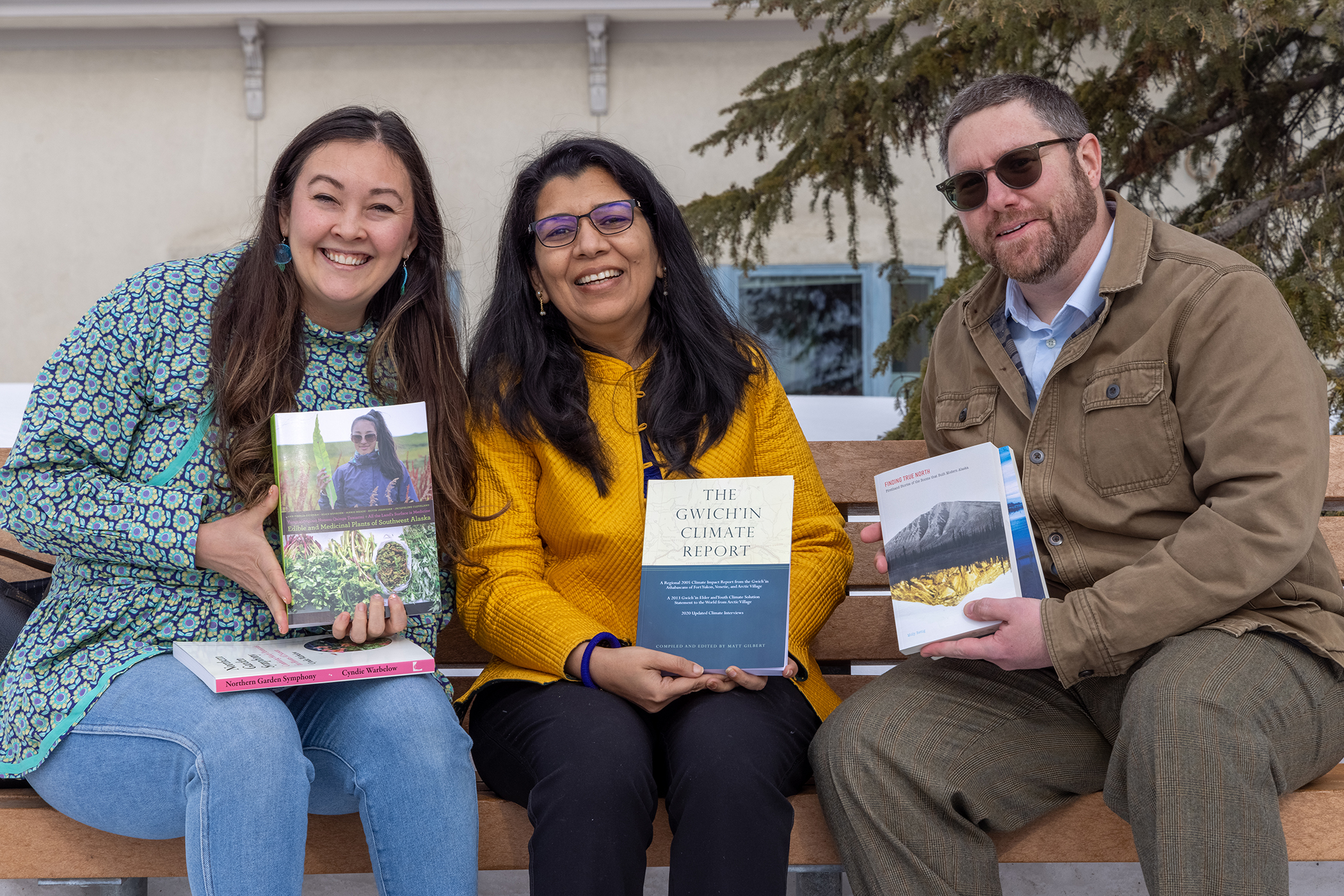 Tia Tidwell, Anupma Prakash, and Nate Bauer are seated on an outdoor bench displaying a variety of recent UA Press book covers.