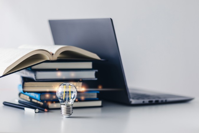 A stack of books, an open laptop computer, writing utensils and a lightbulb sit arranged on a white desktop.