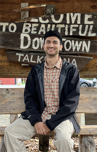 Aaron Salzman, The Sun Star news director, sits on a bench in front of a weathered, hand painted wooden sign that reads 